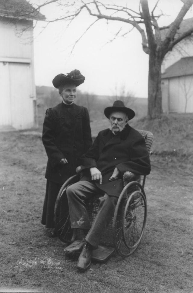 black and white photo of James Moore in wheelchair and wife Irene standing beside taken outside in f